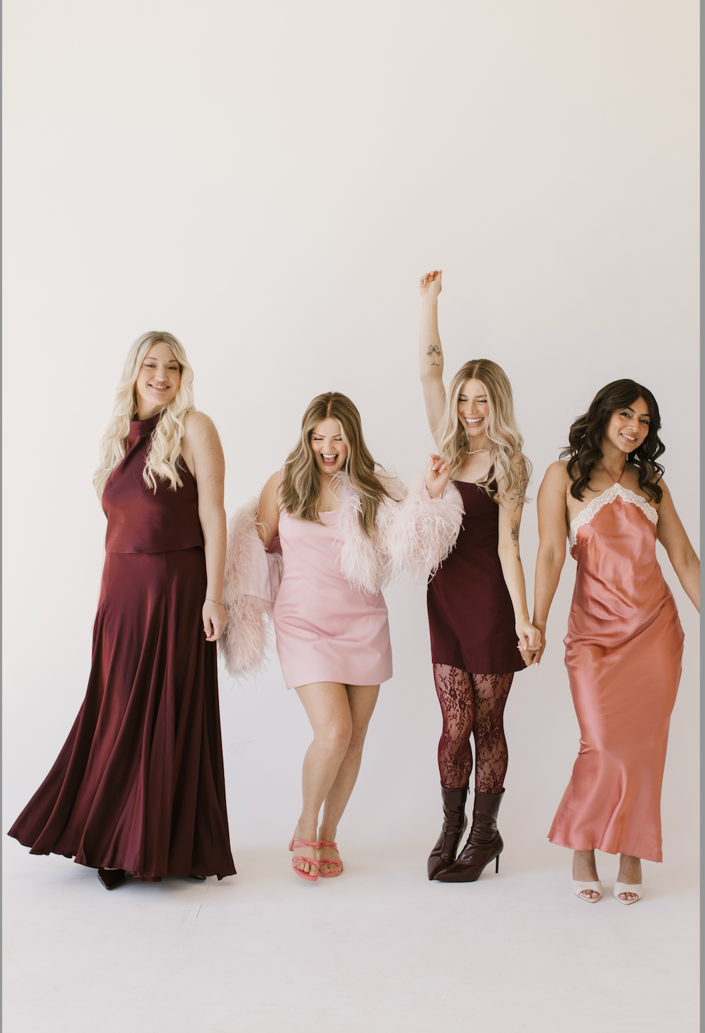 Four women posing in elegant evening gowns against a plain white studio background. The dresses include a floor-length maroon halter gown, a short pink dress with feather trim, a sleeveless maroon dress with lace tights, and a peach satin slip dress.